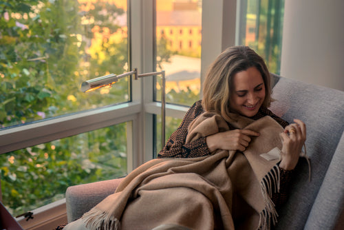 Smiling woman curled in a gray armchair by a sunny window, wrapped in the beige side of the reversible Cielo baby alpaca throw blanket