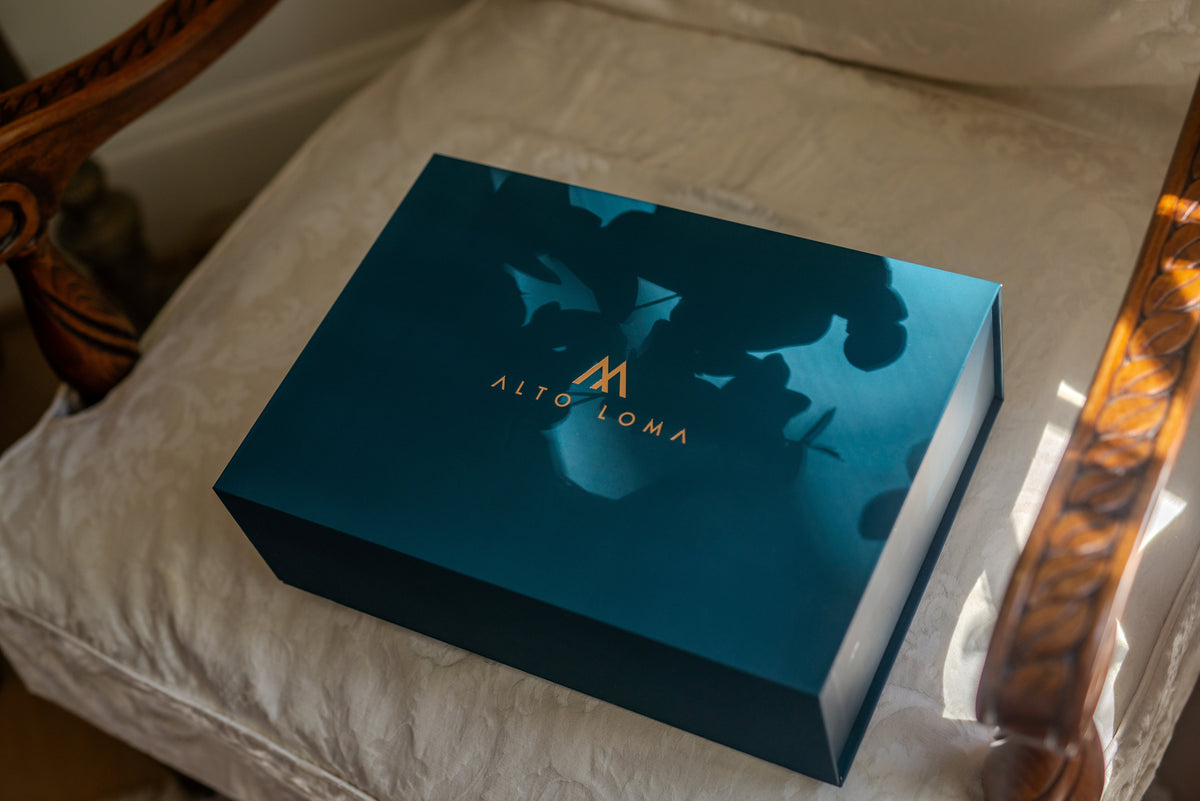 Shadow Teal ALTO LOMA gift box with a copper foil logo, resting on an ivory throw blanket in dappled light