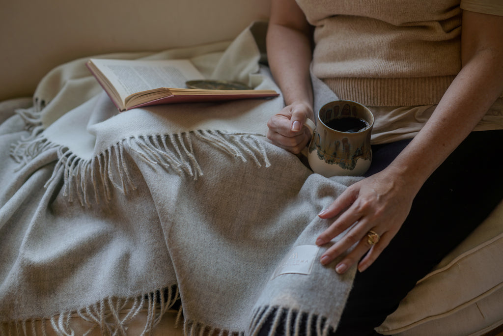 Woman on a sofa with the light gray Sillar baby alpaca throw blanket across her lap, a mug of coffee in one hand and an open book beside her