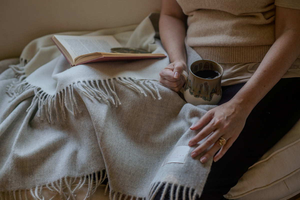 Woman on a sofa with the light gray Sillar baby alpaca throw blanket across her lap, a mug of coffee in one hand and an open book beside her