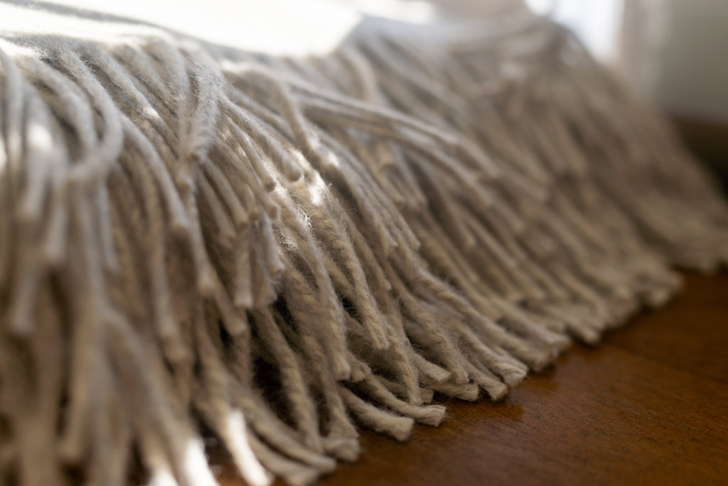 Twisted fringe of the Sillar baby alpaca throw blanket in soft window light, resting on a wooden table
