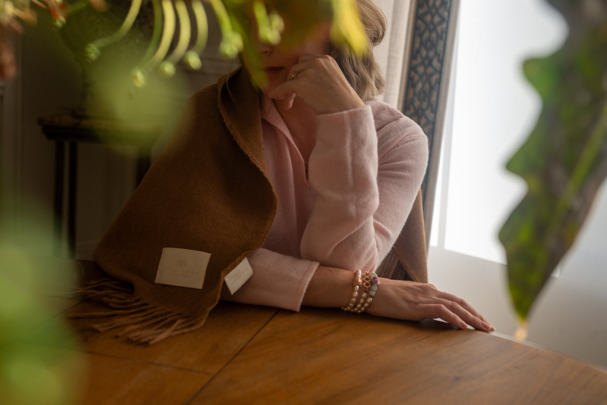 Woman seated at a wooden table with the camel Tero baby alpaca throw blanket draped over her shoulder