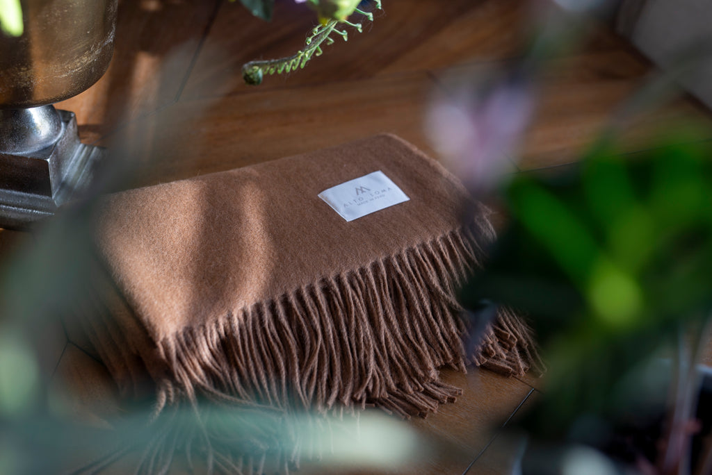 Folded camel Tero baby alpaca throw blanket with ALTO LOMA label on a wooden table among green plants